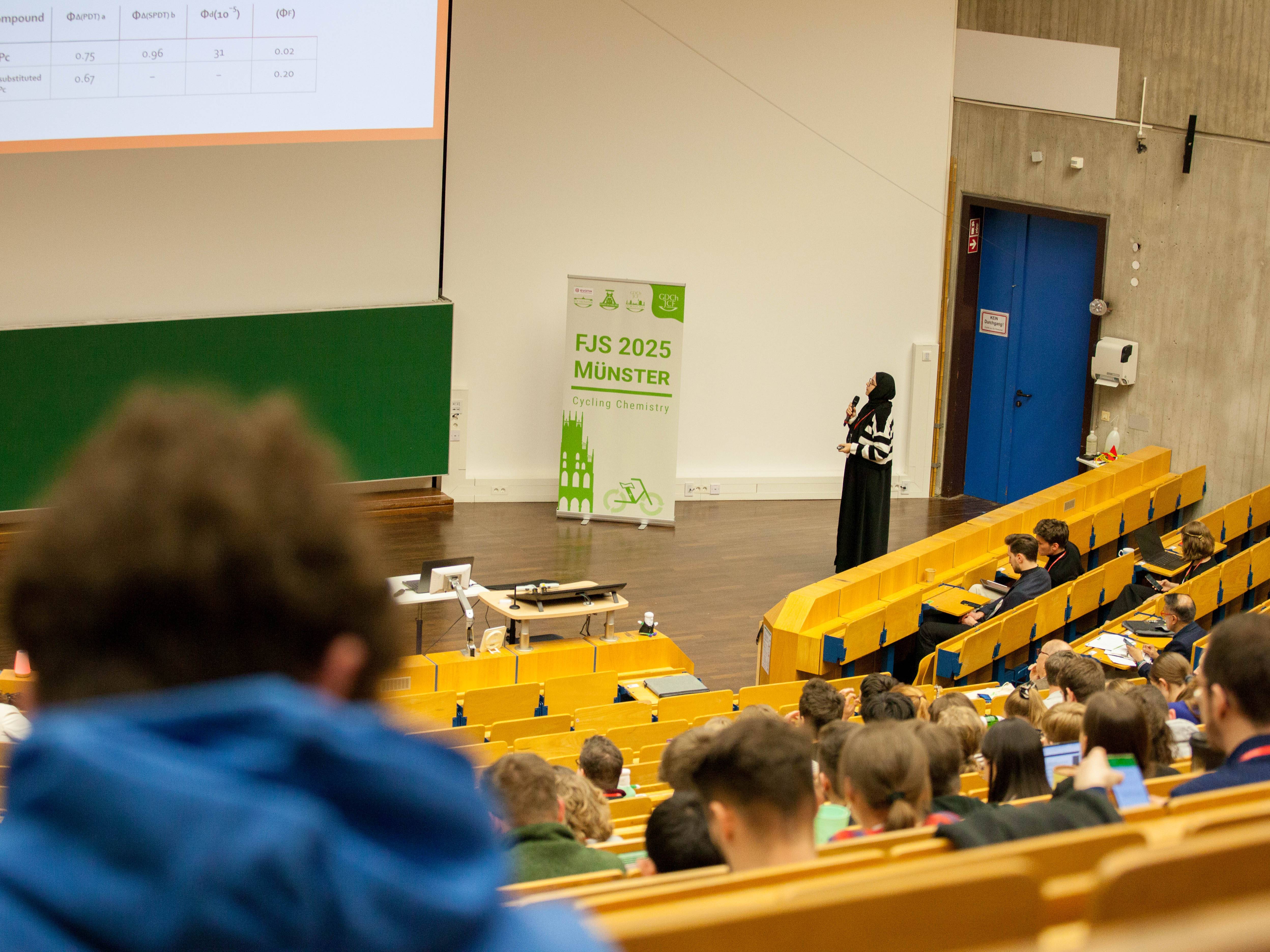 Ein Hörsaal von hinten fotografiert mit vielen im Plenum. Vorne eine junge Frau, die referiert.