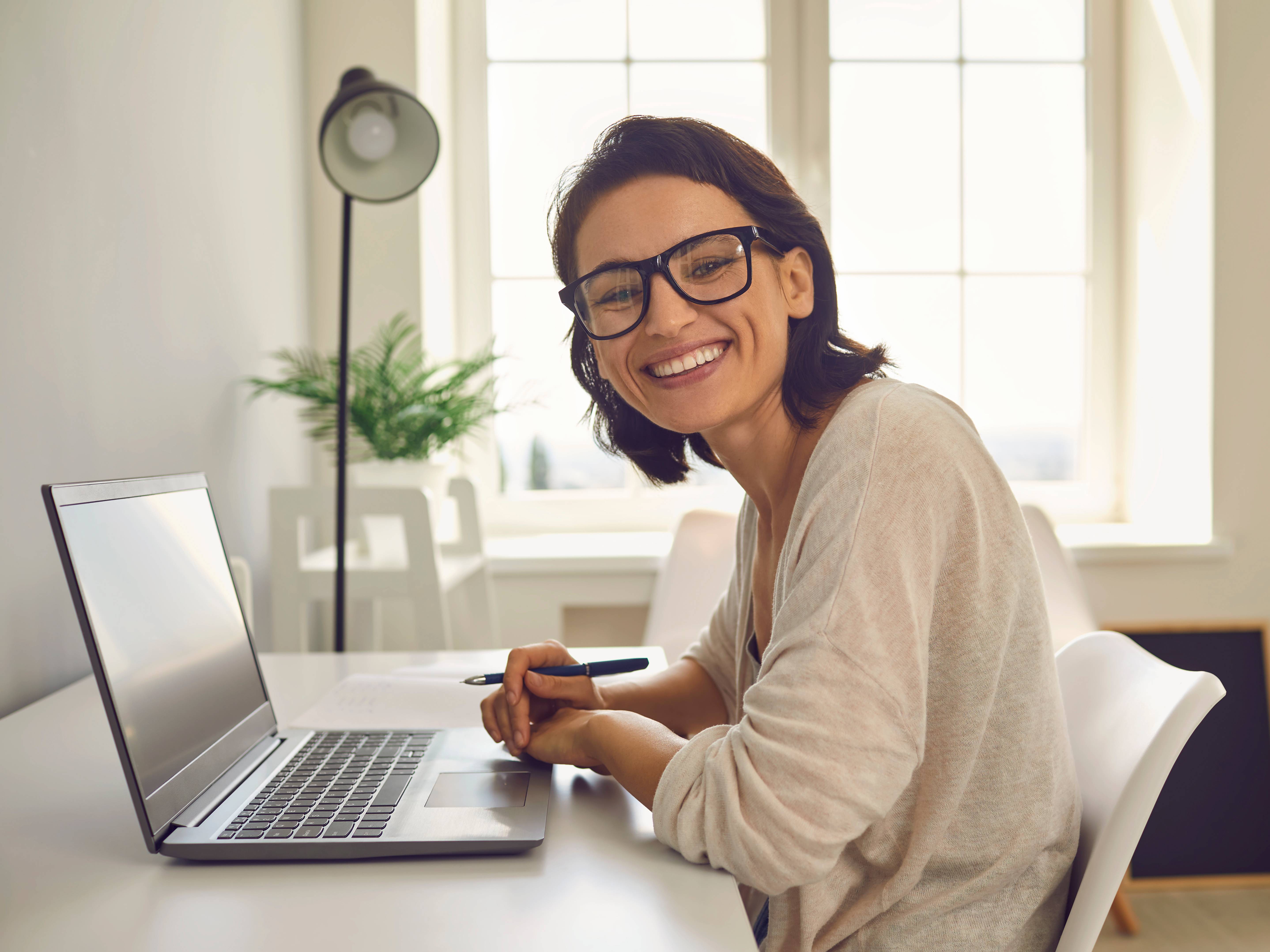 Junge Frau sitzt am Tisch vor ihrem Laptop und lacht in die Kamera