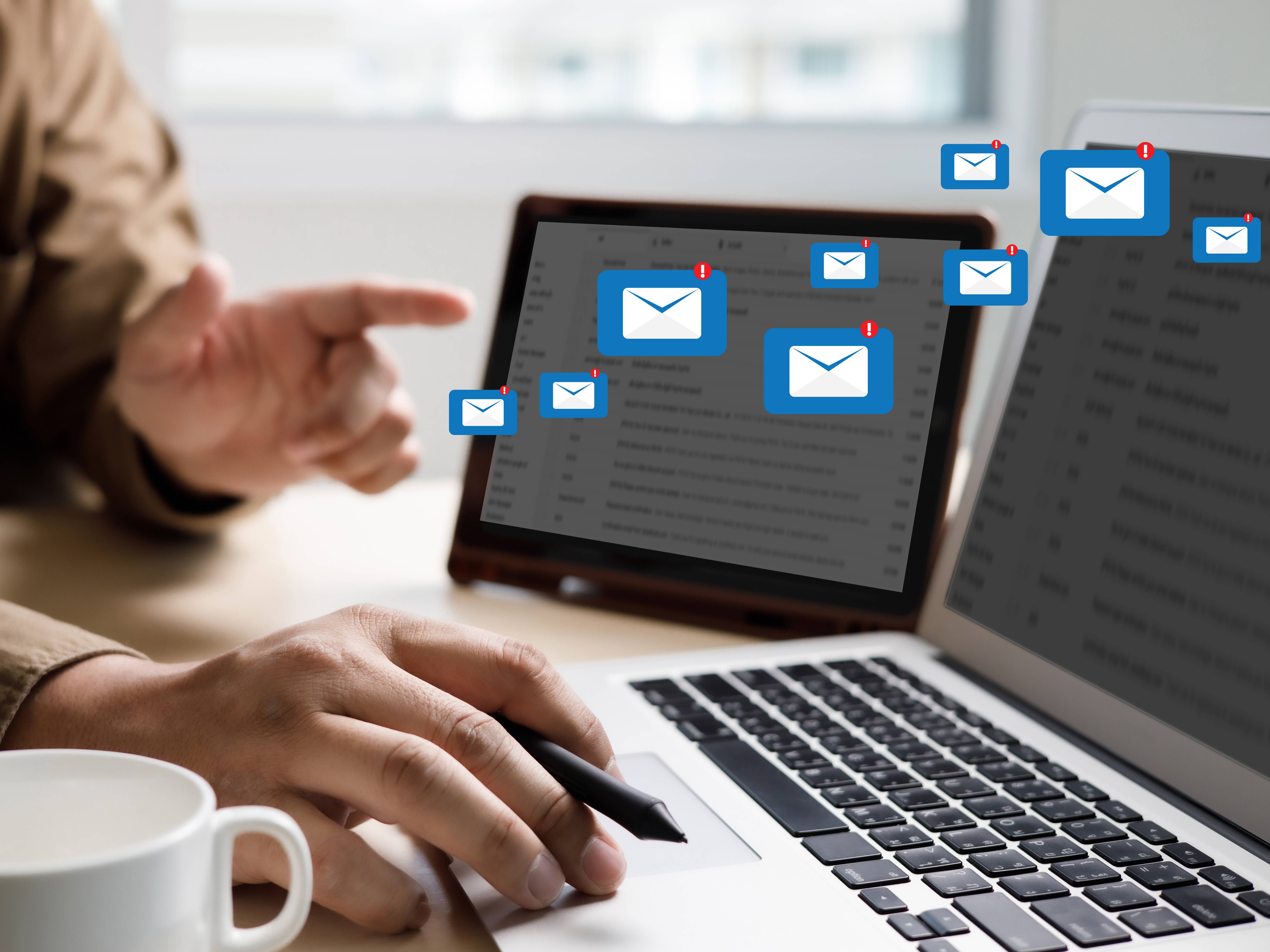 Zwei Laptops nebeneinander. E-Mail Symbole fliegen vom einen Bildschirm zum anderen. Davor jemand, der auf den rechten Bildschirm zeigt.