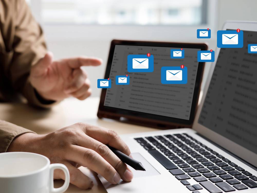 Zwei Laptops nebeneinander. E-Mail Symbole fliegen vom einen Bildschirm zum anderen. Davor jemand, der auf den rechten Bildschirm zeigt.