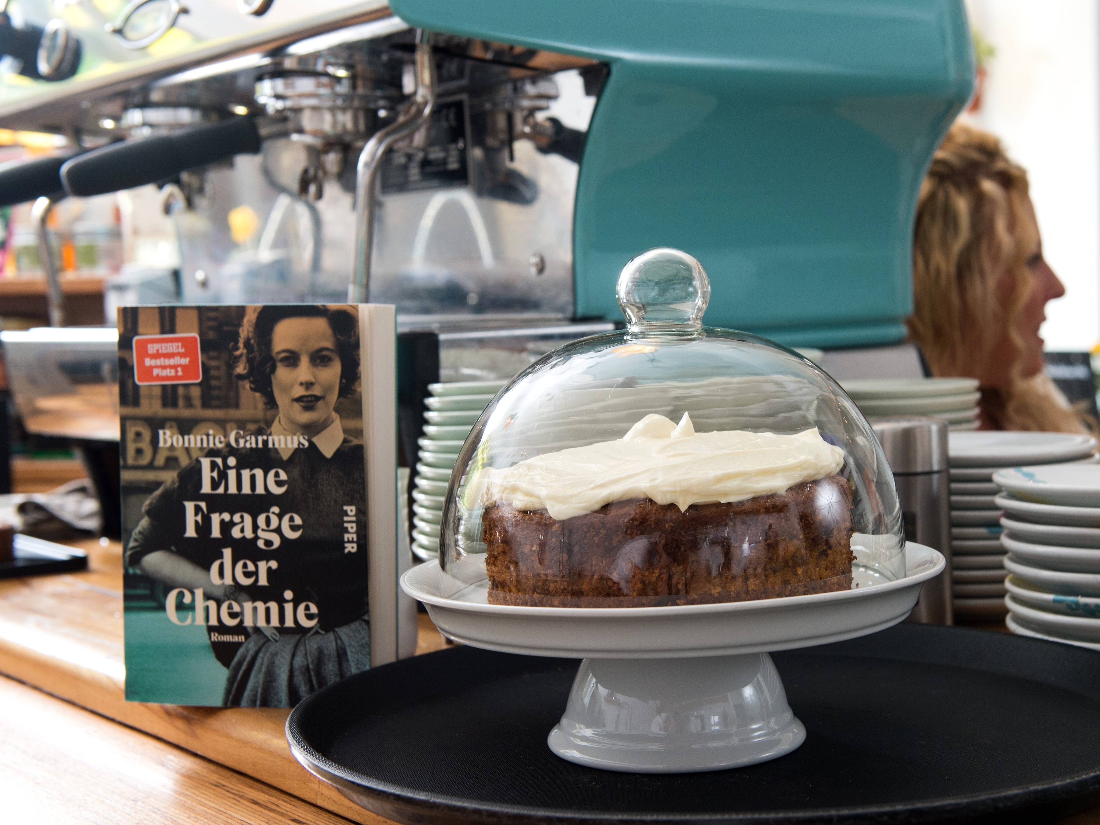 Blick hinter den Tresen des Cafés Don Melone in Düsseldorf. Zu sehen ist ein selbstgebackener Kuchen unter einer Kuchenglocke und daneben ein Buch zum Backen mit dem Titel „Eine Frage der Chemie“.