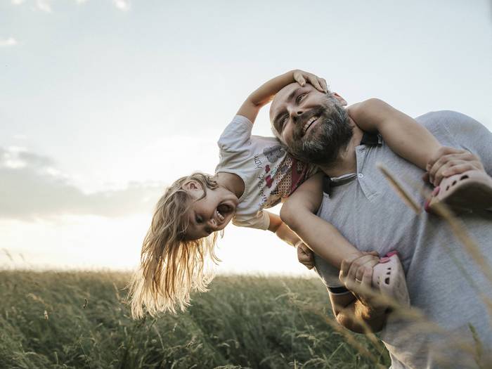 Mann im Feld beugt sich von rechts ins Bild. Kind sitzt auf seinen Schultern. Beide lachen. Sonne im Hintergrund.