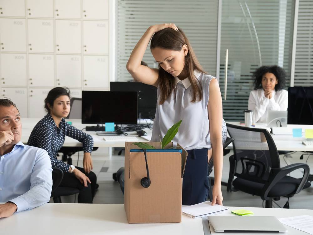 Eine Frau ist gekündigt worden und steht traurig am Tisch in einem Großraumbüro. Vor ihr steht ein Karton mit ihren persönlichen Sachen. Die Kolleginnen und Kollegen neben und hinter ihr schauen sie mitleidig an.