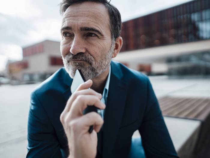 Nahaufnahme von einem älteren Mann, der in die Ferne schaut, mit seinem Handy in der Hand.