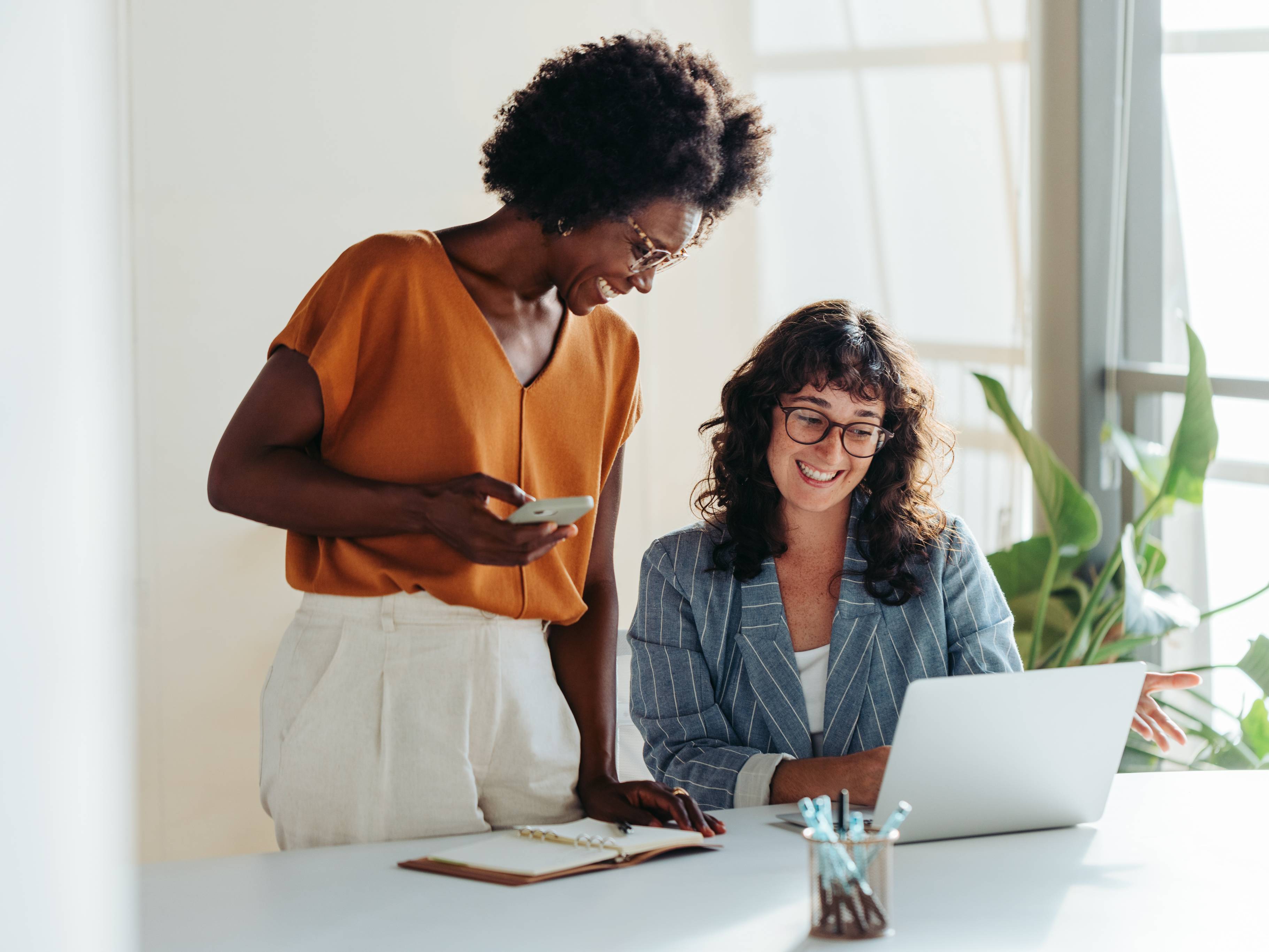Symbolbild: zwei gut gelaunte Frauen an einem Bürotisch, eine sitzend vor dem Laptop, die andere stehend.